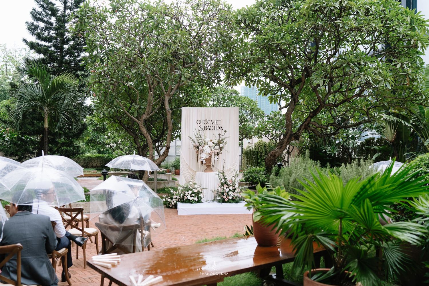 Outdoor wedding ceremony in a garden with guests holding clear umbrellas in the rain near a floral altar.