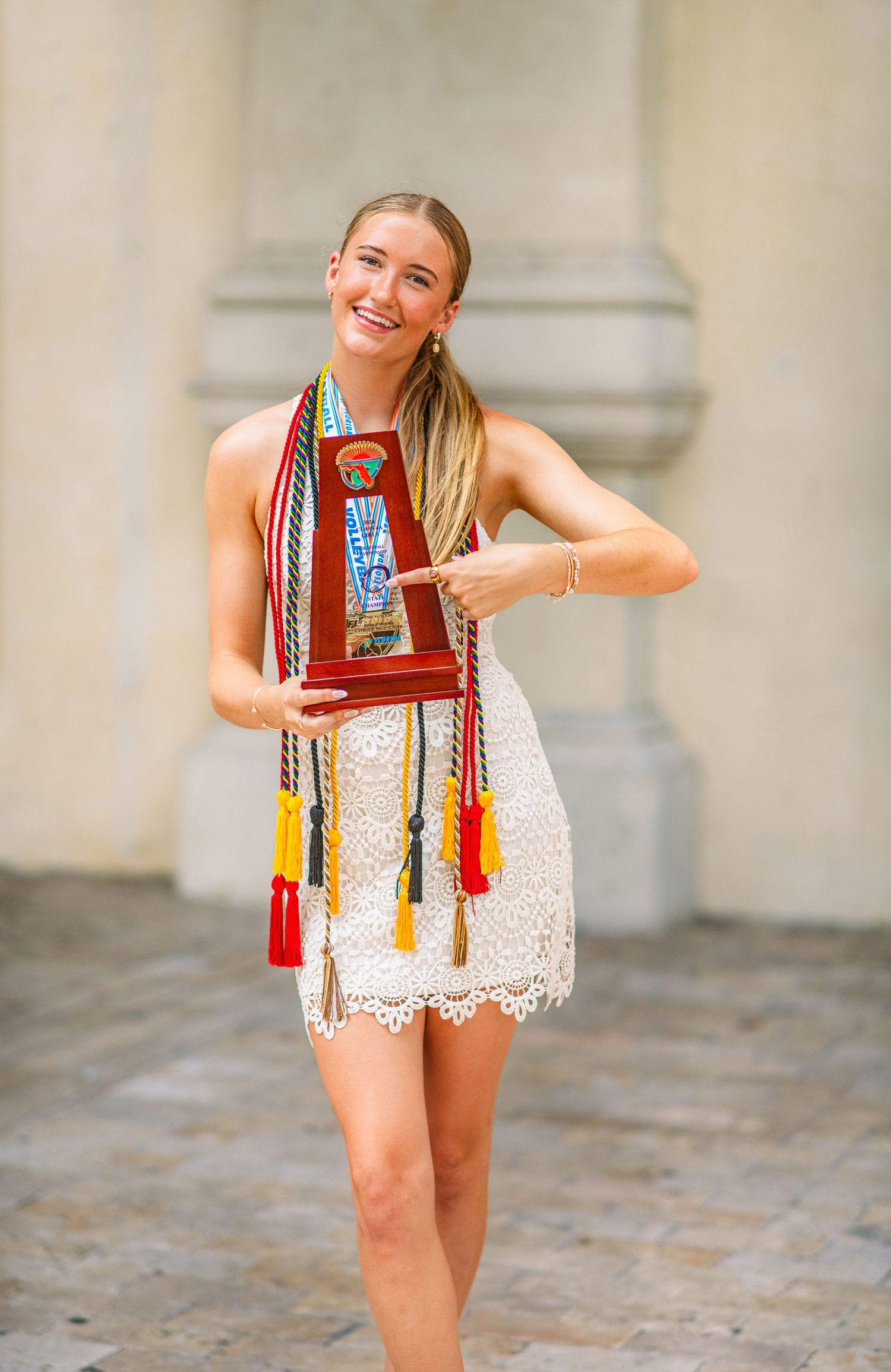 Smiling graduate in white lace dress holding trophy with colorful honor cords at outdoor ceremony.
