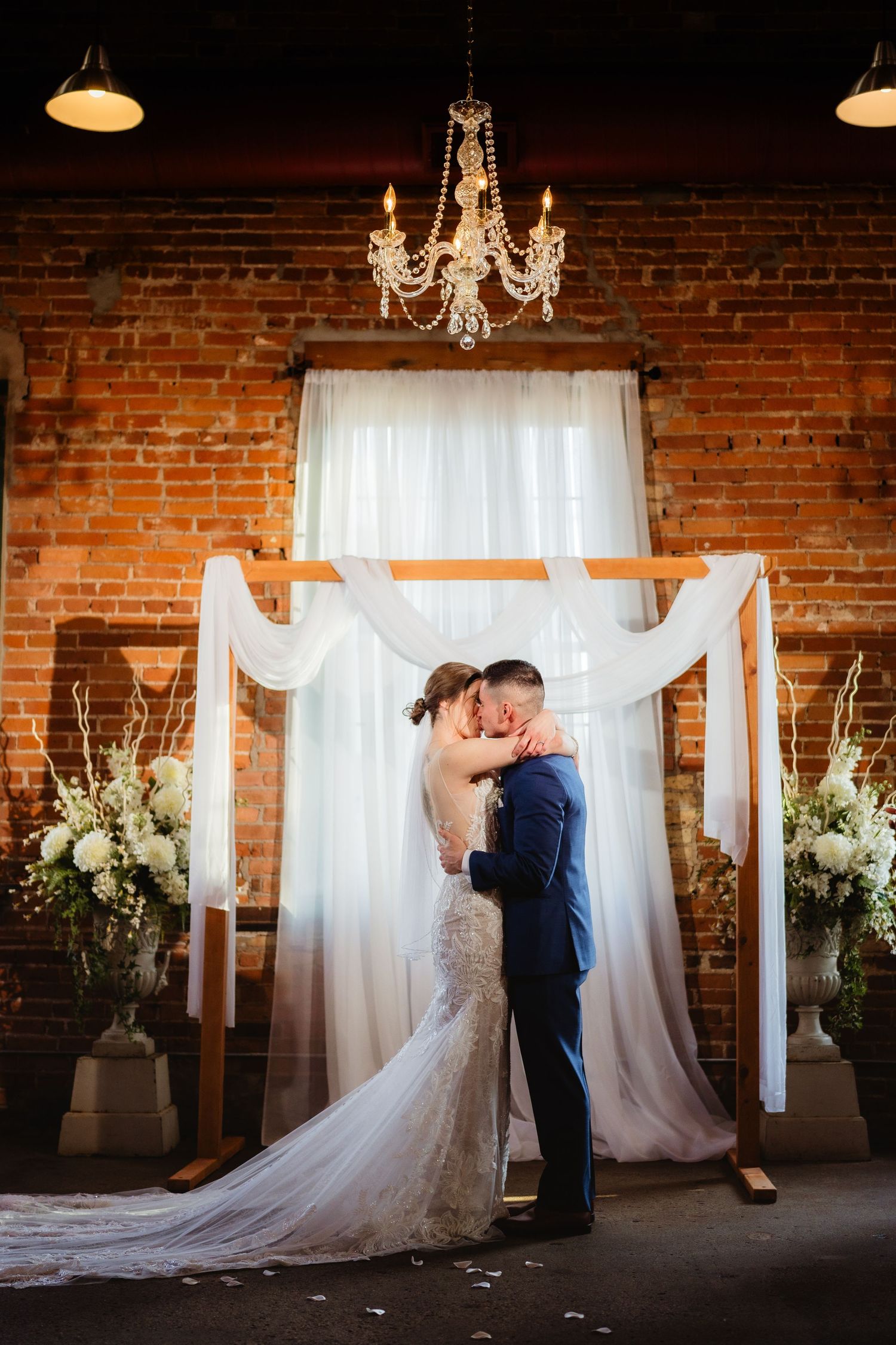 Bride and groom share first kiss under elegant floral arch with white draping in rustic brick wedding venue.