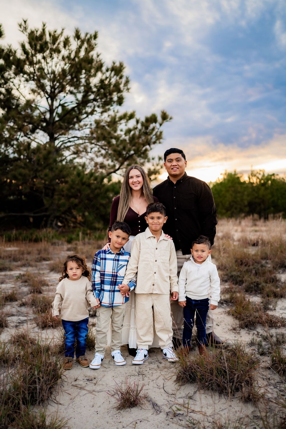 Family of six poses outdoors at sunset on sandy ground with trees and colorful sky in background.