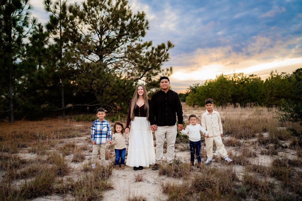 Family of six poses at sunset in a pine forest clearing, parents holding hands with four young children on a sandy path.
