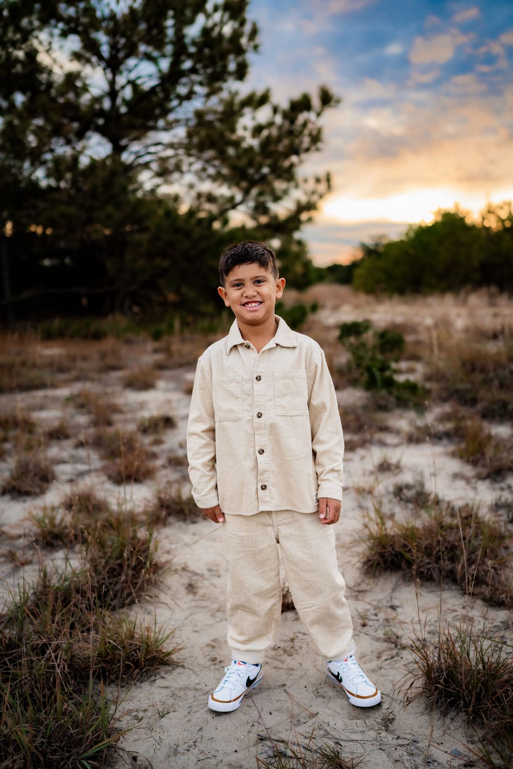 A smiling boy in a beige linen outfit and Nike sneakers stands on sandy ground at sunset with trees behind him.
