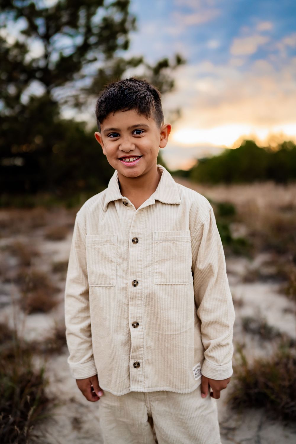 A smiling young boy in a beige linen shirt stands outdoors at sunset with trees and colorful sky behind him.