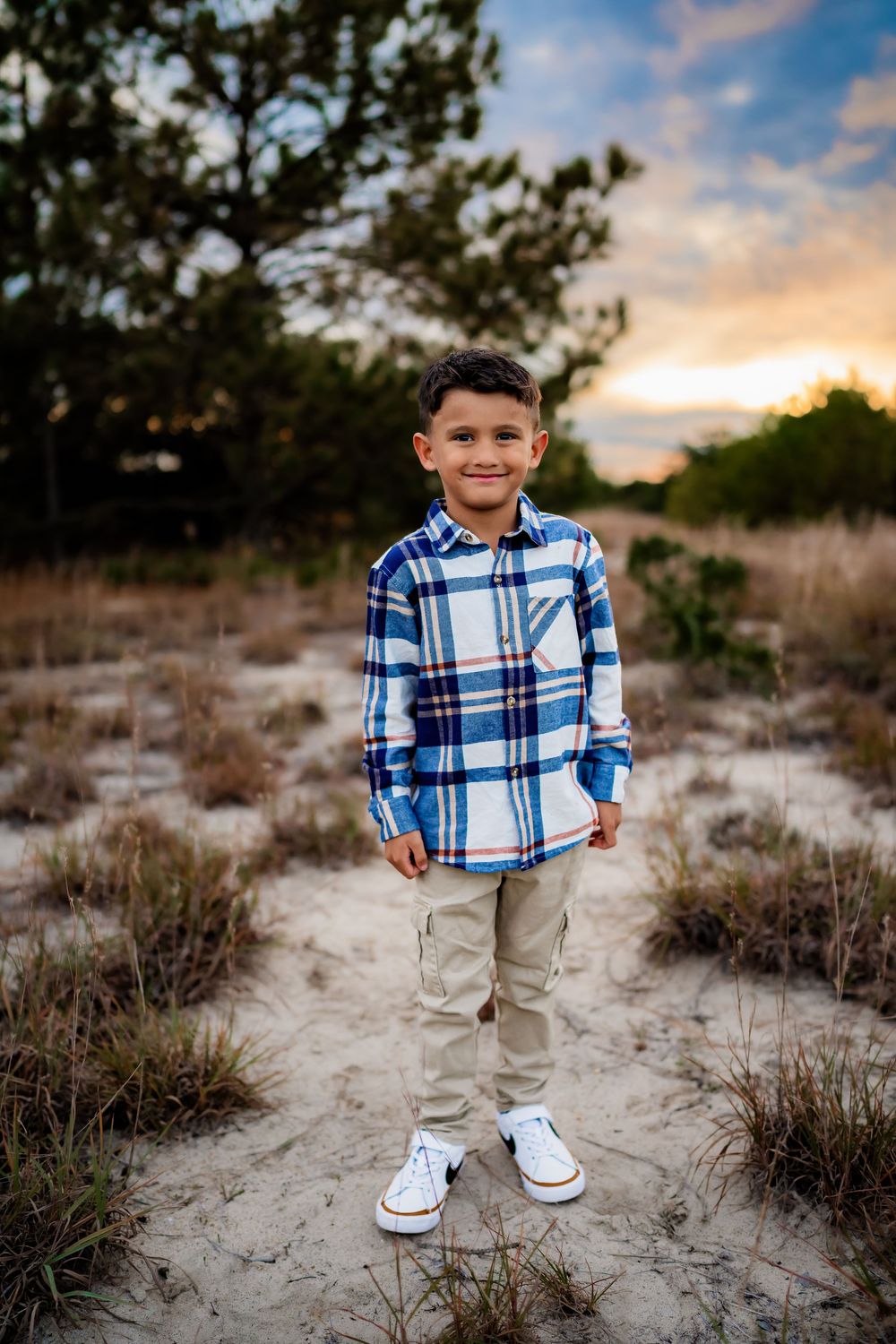 Young boy in plaid shirt and khaki pants smiling at sunset on sandy beach path.