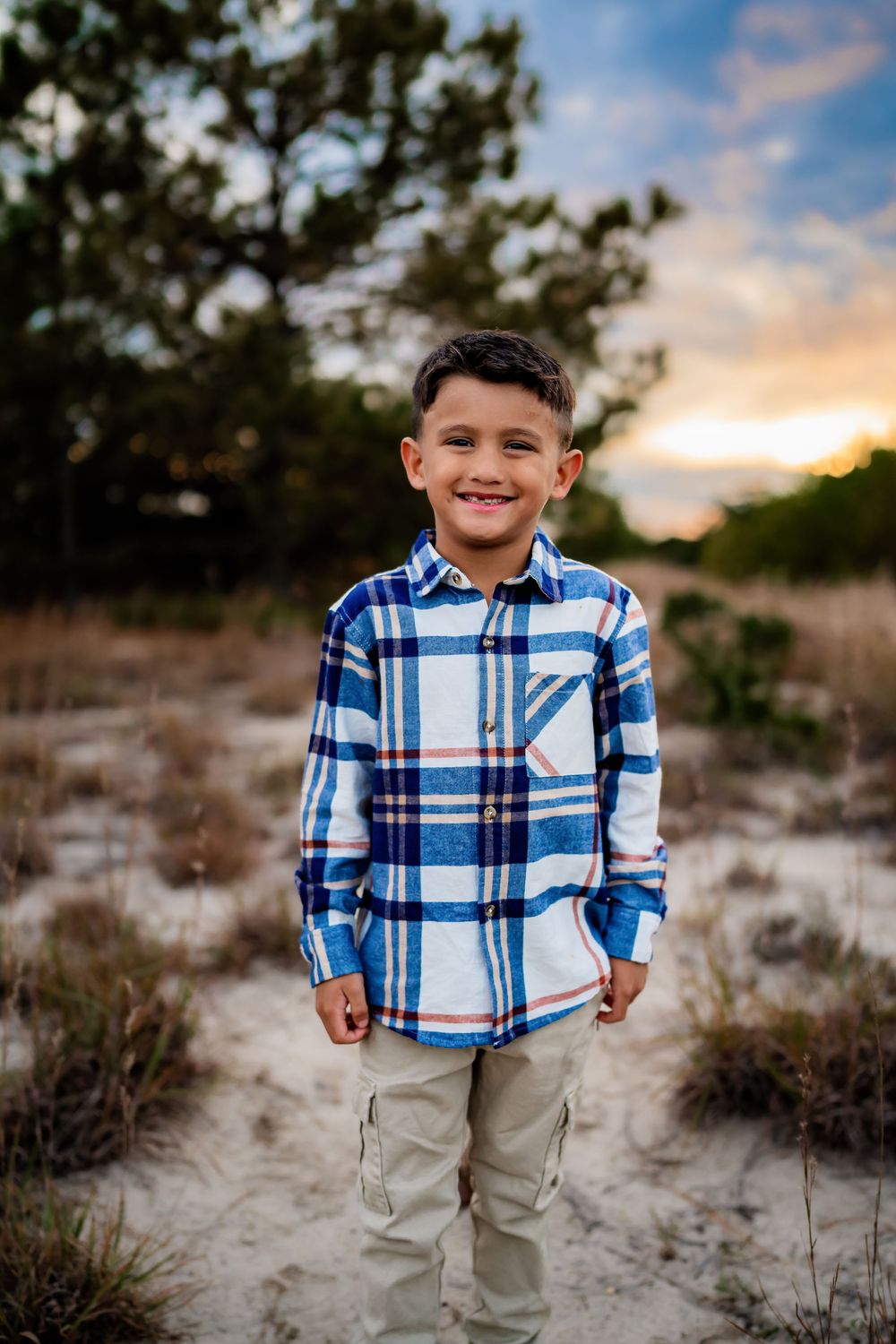 Smiling young boy in blue plaid shirt and khaki pants standing on sandy path at sunset.