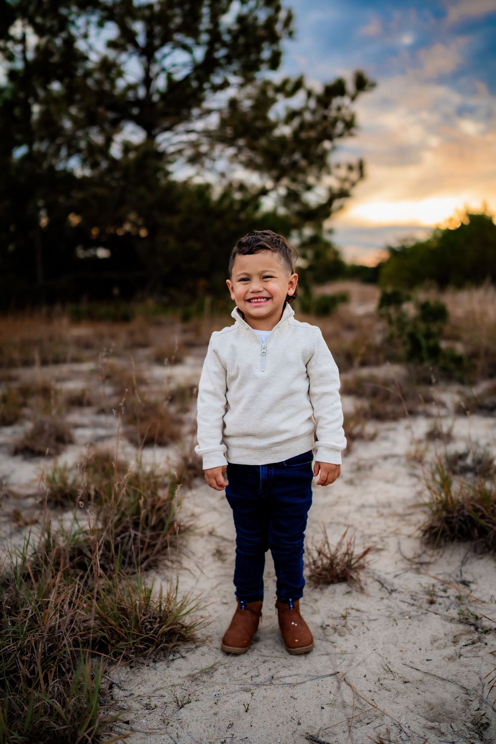 Smiling young boy in white sweater and jeans stands on sandy path at sunset.