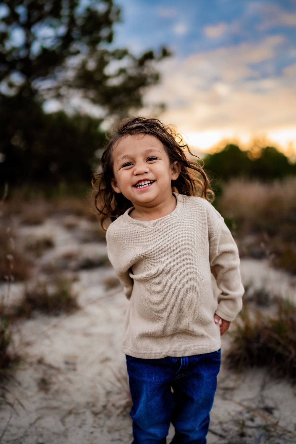Smiling toddler with curly hair in beige sweater and jeans standing on sandy path at sunset.