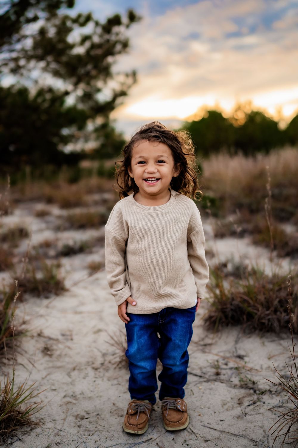 Smiling young child with curly hair in beige sweater and jeans standing on sandy path at sunset.