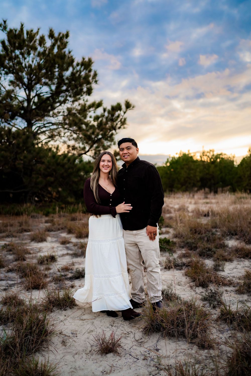 Couple poses together on sandy ground at sunset with pine trees and colorful sky behind them.