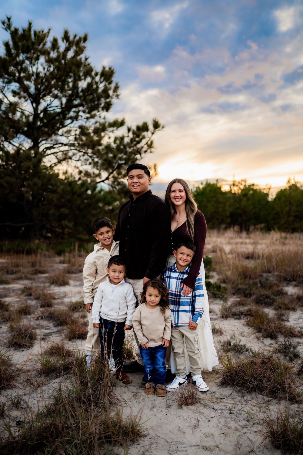 Family of six poses outdoors on sandy terrain at sunset with pine trees and dramatic sky.