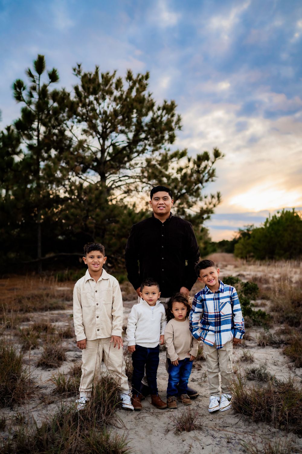 Father poses with three young sons on sandy trail surrounded by trees at sunset.
