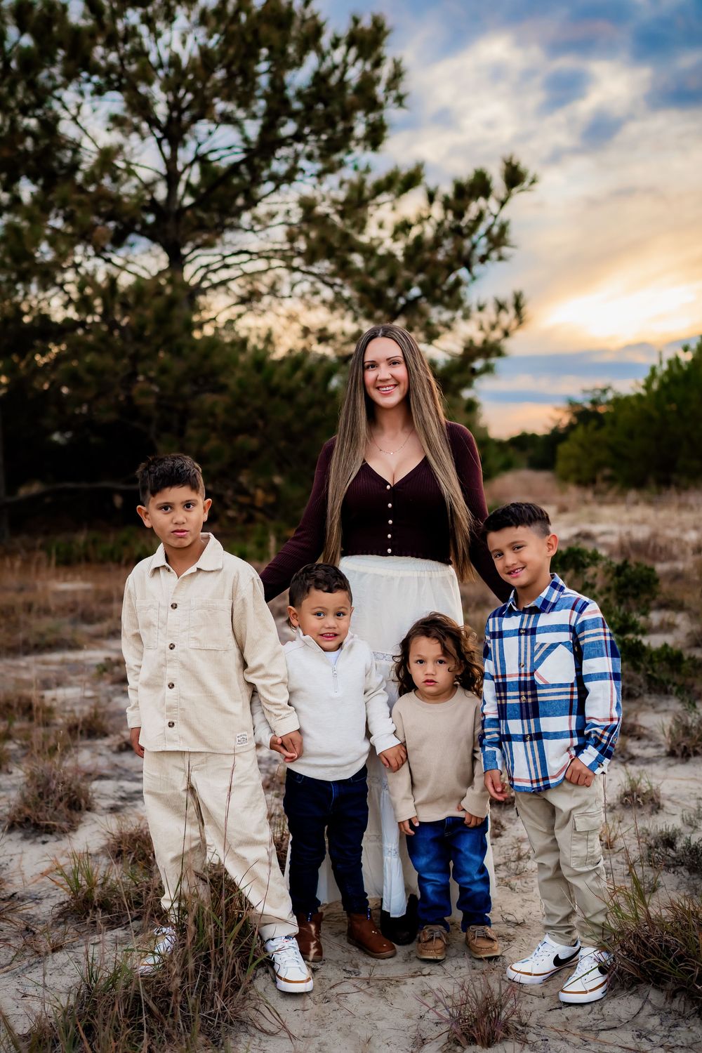 A woman with long hair stands smiling with four young boys in a sandy outdoor setting at sunset.