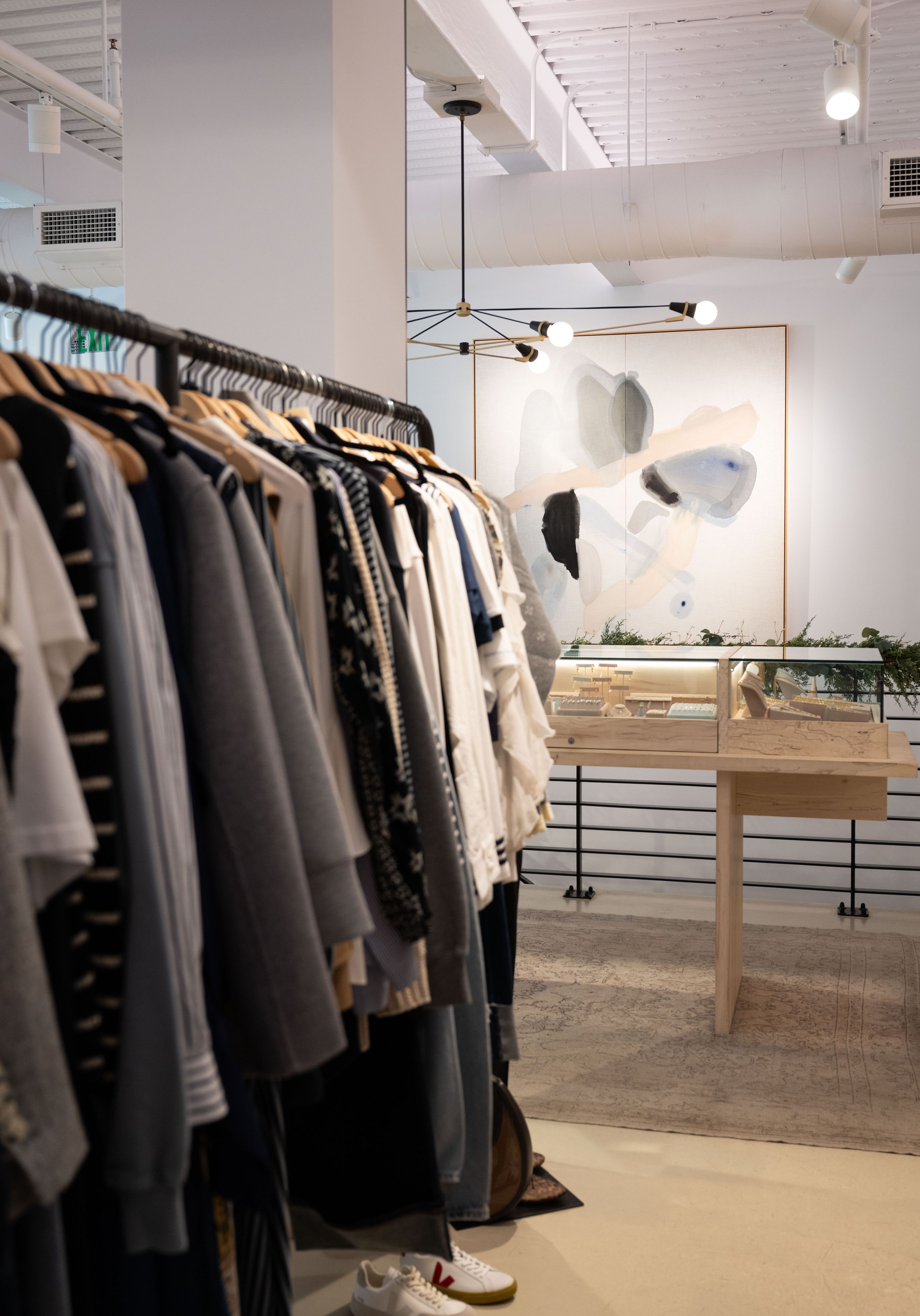 Modern boutique clothing store interior with garment racks, neutral tones, abstract art, and wooden display tables.