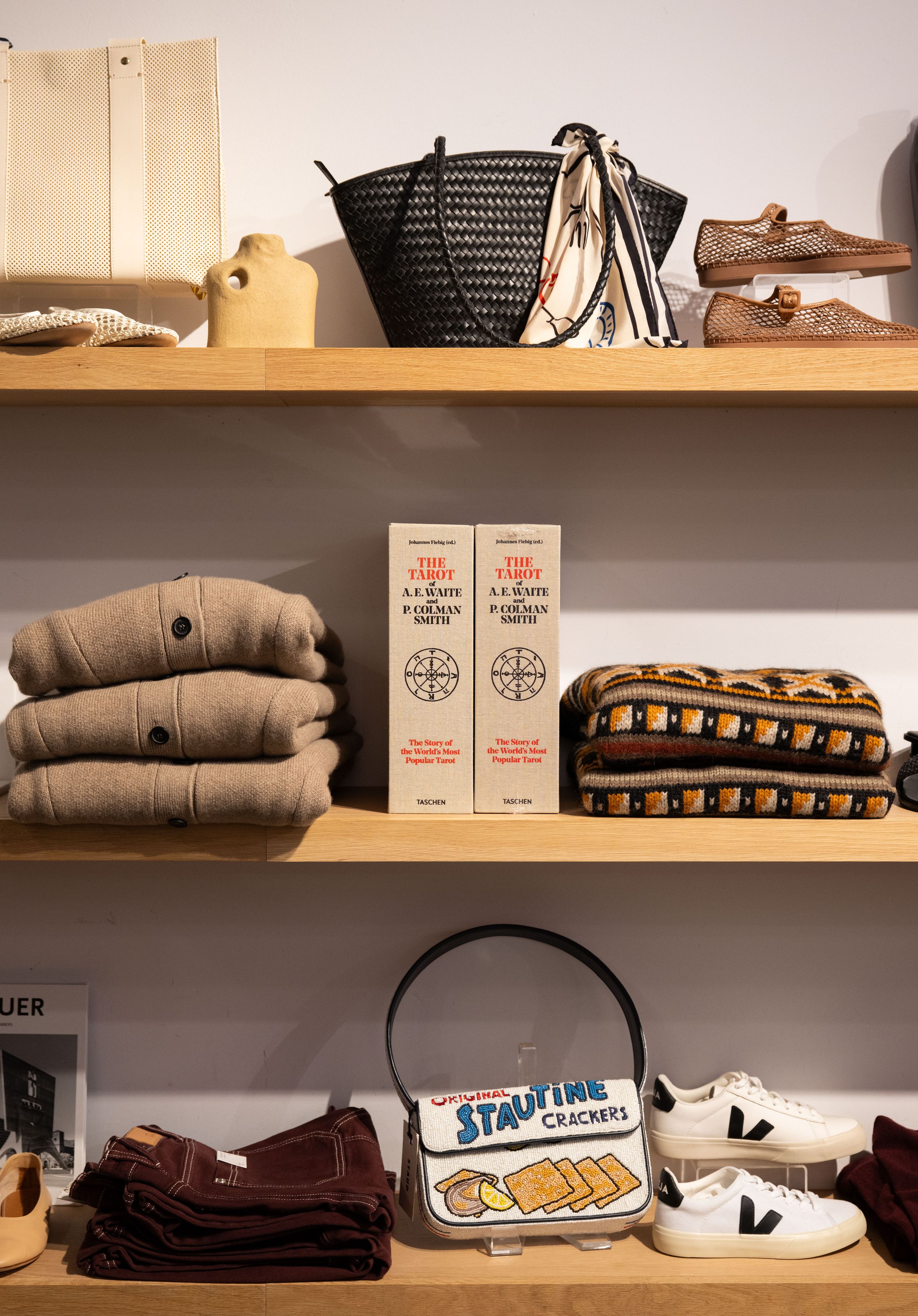 Retail store shelves displaying folded sweaters, woven bags, white sneakers, a crossbody bag, and books in a boutique setting.