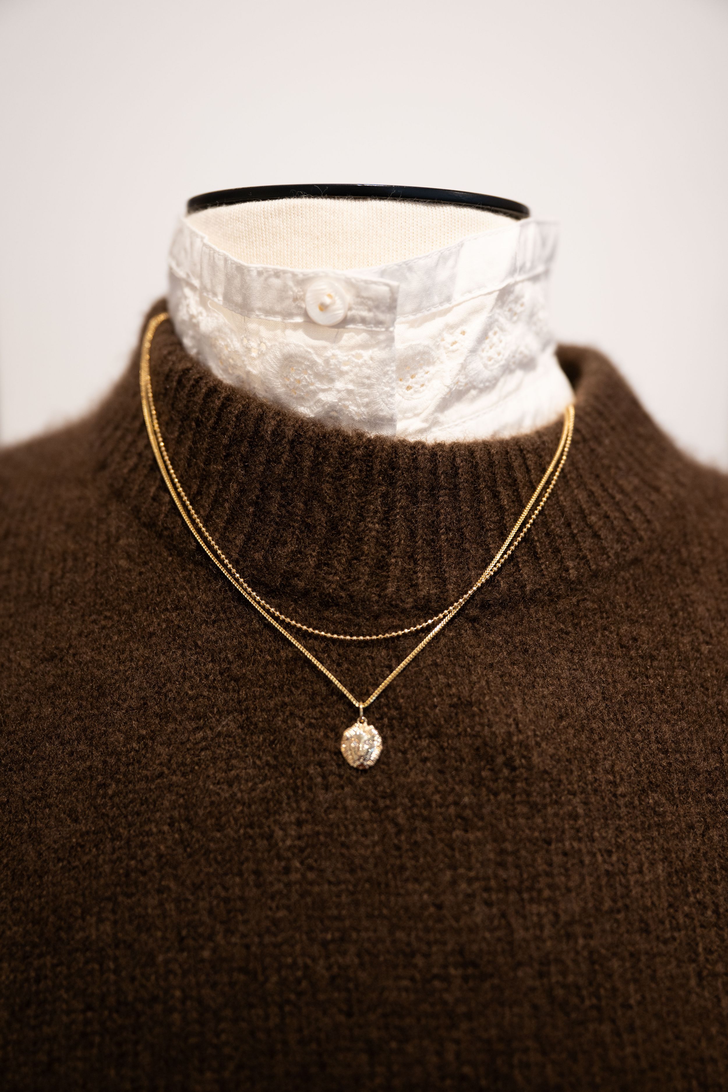 Gold layered necklace with pendant on brown knit turtleneck sweater over white lace collar on mannequin.