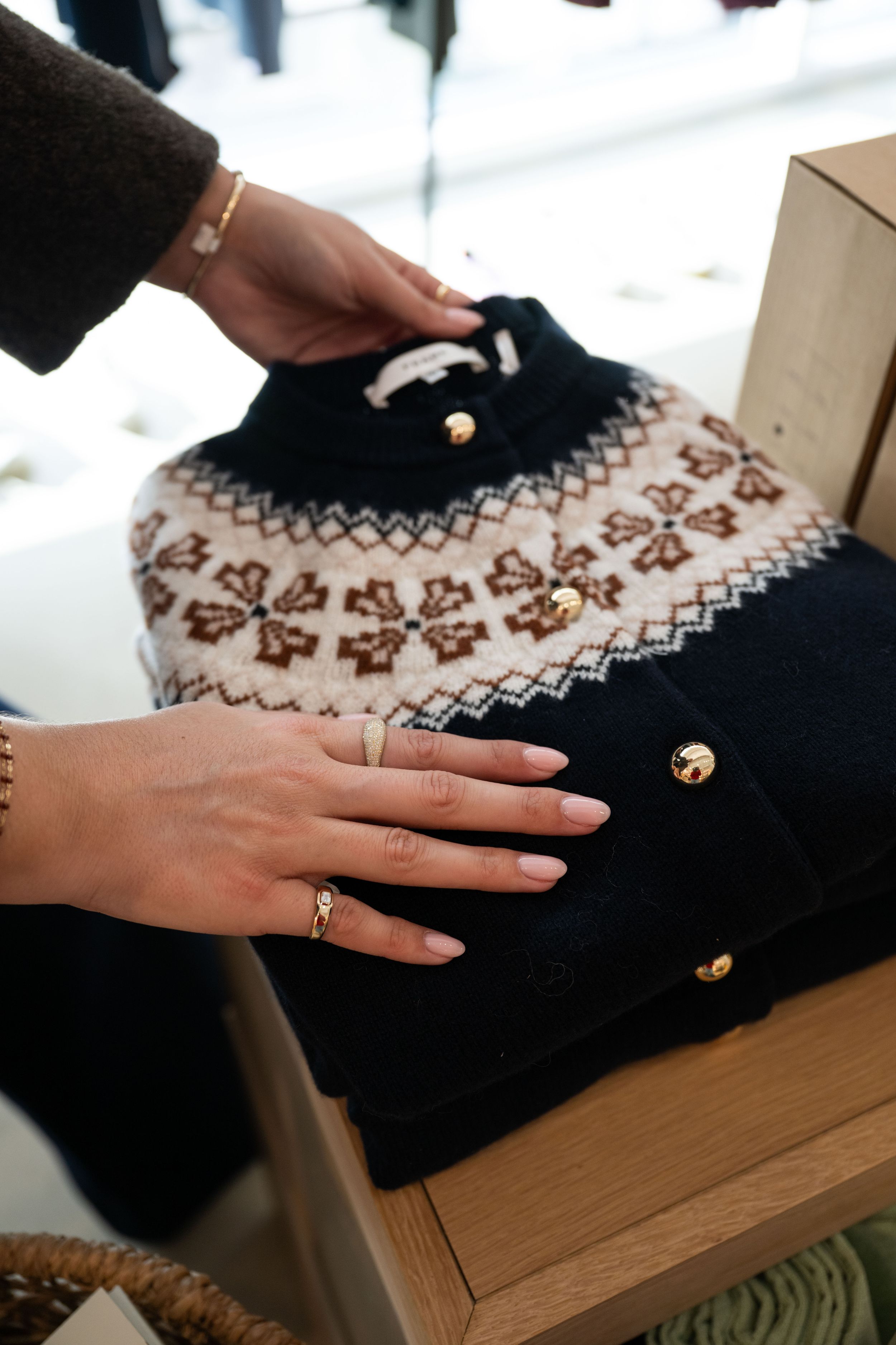 Hands arranging a folded Nordic fair isle sweater with gold buttons on a wooden shelf in a boutique.