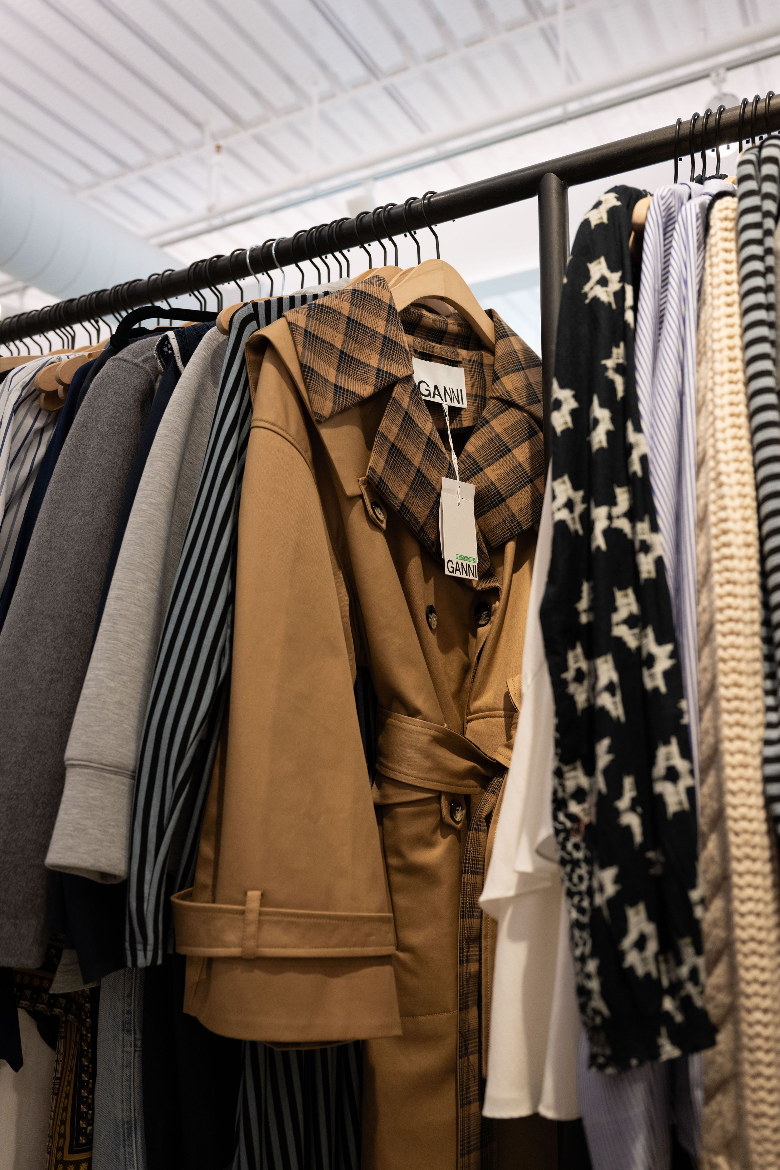 GANNI clothing rack displaying a tan trench coat with plaid collar, striped and patterned garments in a boutique store.