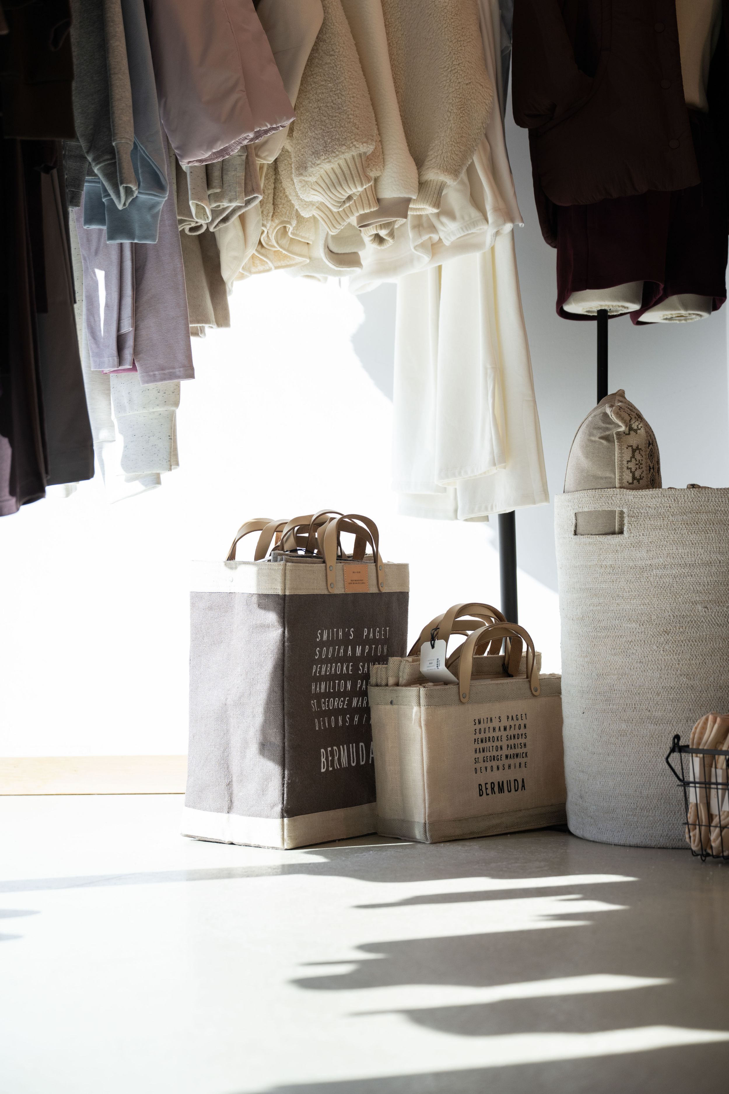 Boutique clothing rack with hanging garments and Berwood canvas tote bags on a sunlit white floor.