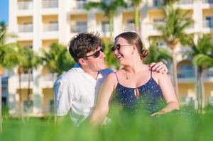 A happy couple in sunglasses smiling at each other on a tropical resort lawn with palm trees and a hotel in the background.