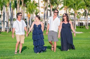 Two couples walk hand-in-hand on lush green grass surrounded by tall palm trees in a tropical resort setting.