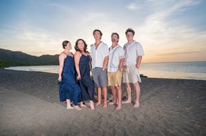 Family of five posing together on a dark sand beach at sunset with tropical hills in the background.