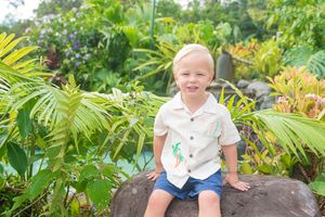 A small figure sits on a rock surrounded by lush tropical foliage and palm fronds in a garden setting.