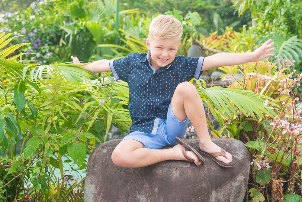 A young person sits on a large rock surrounded by lush tropical plants and ferns in a garden setting.
