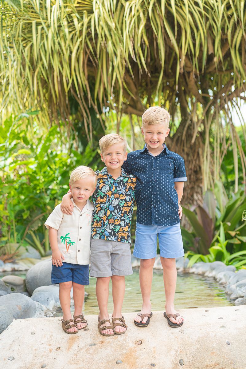 Three children standing together on a sandy beach path with tropical plants in the background.