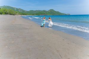 Two brides in flowing gowns hold hands running along a tropical sandy beach with turquoise ocean waves.