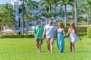 Family of four walking hand-in-hand on lush green lawn at tropical resort with palm trees and white building.