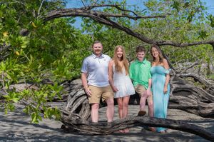 Family of four posing among driftwood and mangroves on a tropical beach, smiling in casual summer outfits.