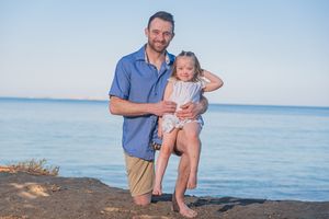 A parent and child stand together on a rocky beach during a sunny day with calm blue water in the background.