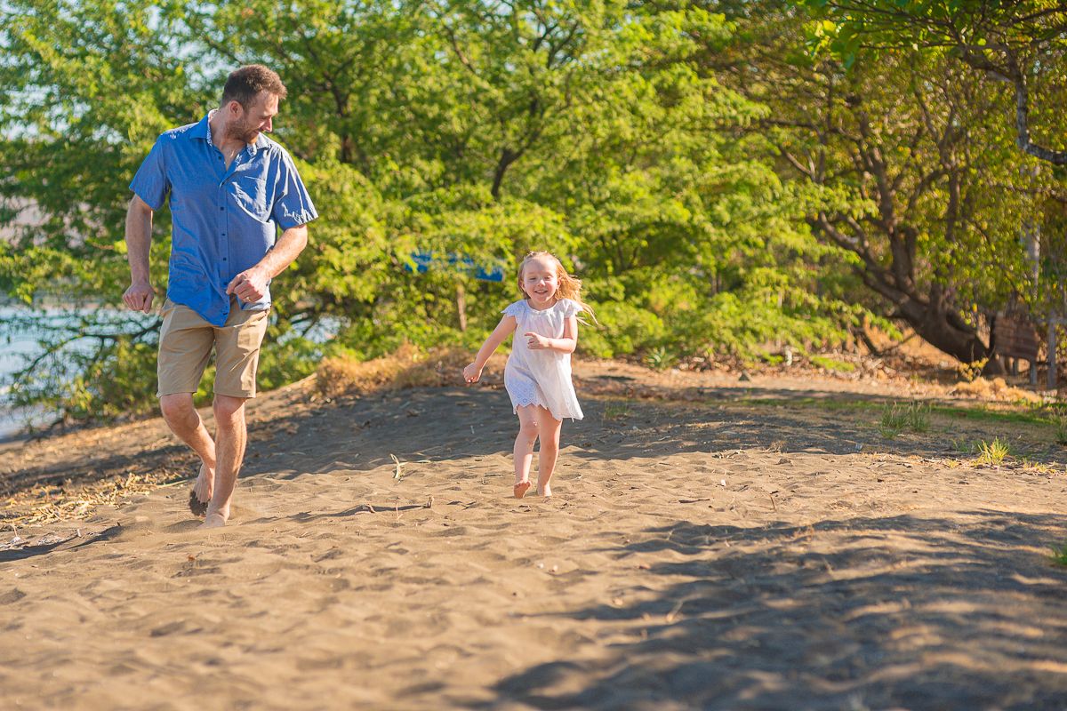 Two people running and playing together on a sandy beach with lush green trees in the background.