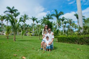 Family of four posing on lush green lawn surrounded by tall palm trees under a bright blue sky.