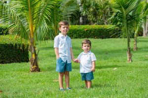 Two young boys in matching white shirts and blue shorts hold hands on a lush green lawn with tropical palm trees.