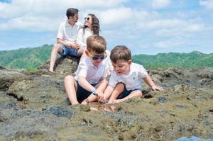 Two boys explore rocky tide pools while parents sit on rocks with lush green hills and cloudy sky behind them.