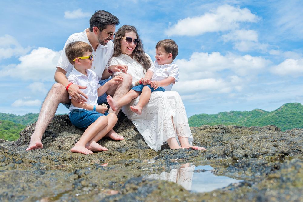 Family of five sitting on coastal rocks with lush green hills and blue sky backdrop on a sunny day.