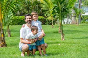 A smiling father crouches with his two young sons on lush green grass surrounded by tropical palm trees.
