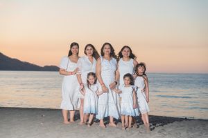 A group in white dresses poses together on a beach at sunset with mountains and ocean in the background.