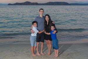 A family stands together on a beach at sunset with mountains and blue water in the background.