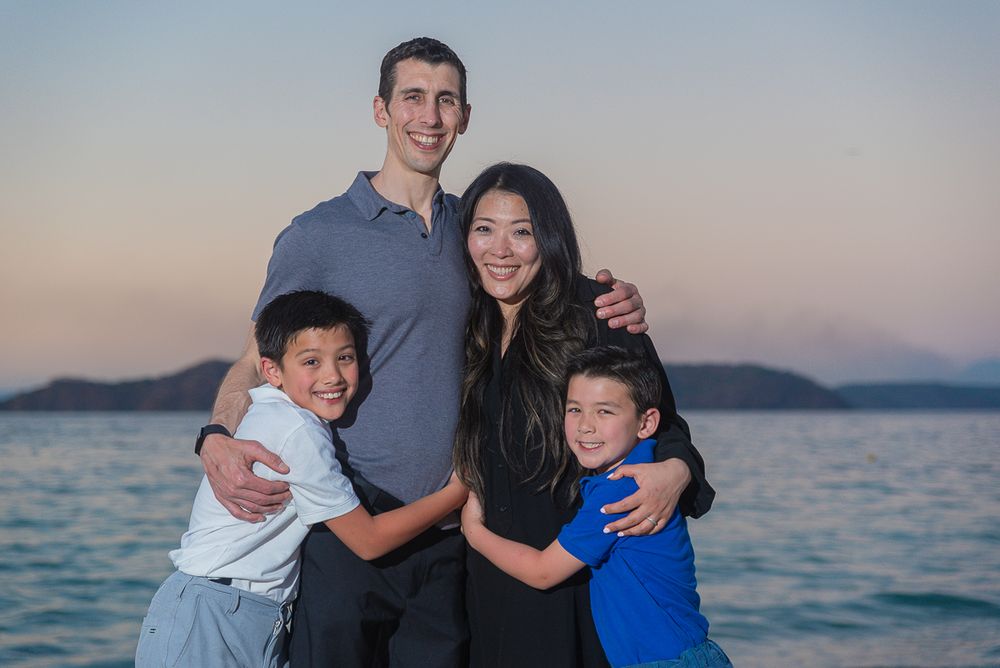 A family of four poses together for a portrait at sunset with an ocean view in the background.
