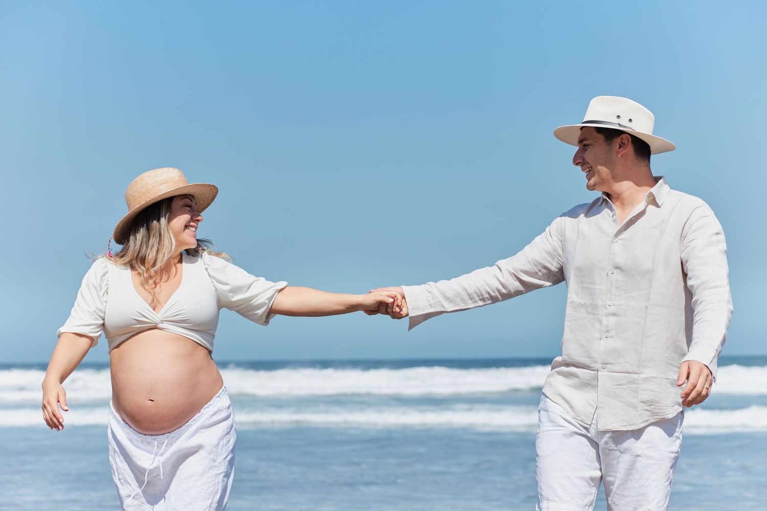 Expecting couple in white outfits holding hands on sunny beach with ocean waves in background.