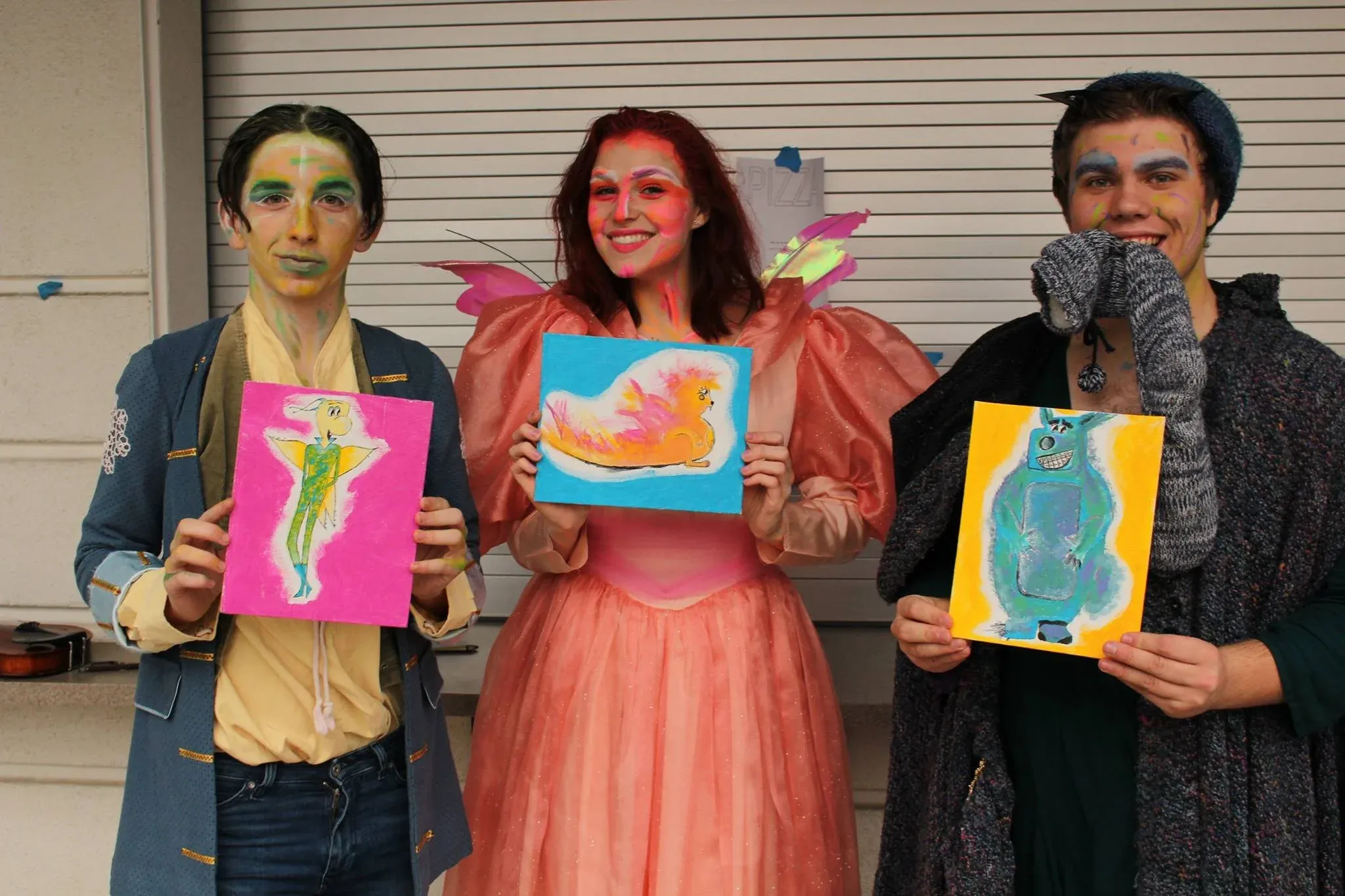 Three performers in costumes hold colorful artwork paintings during a theatrical production or drama class showcase.