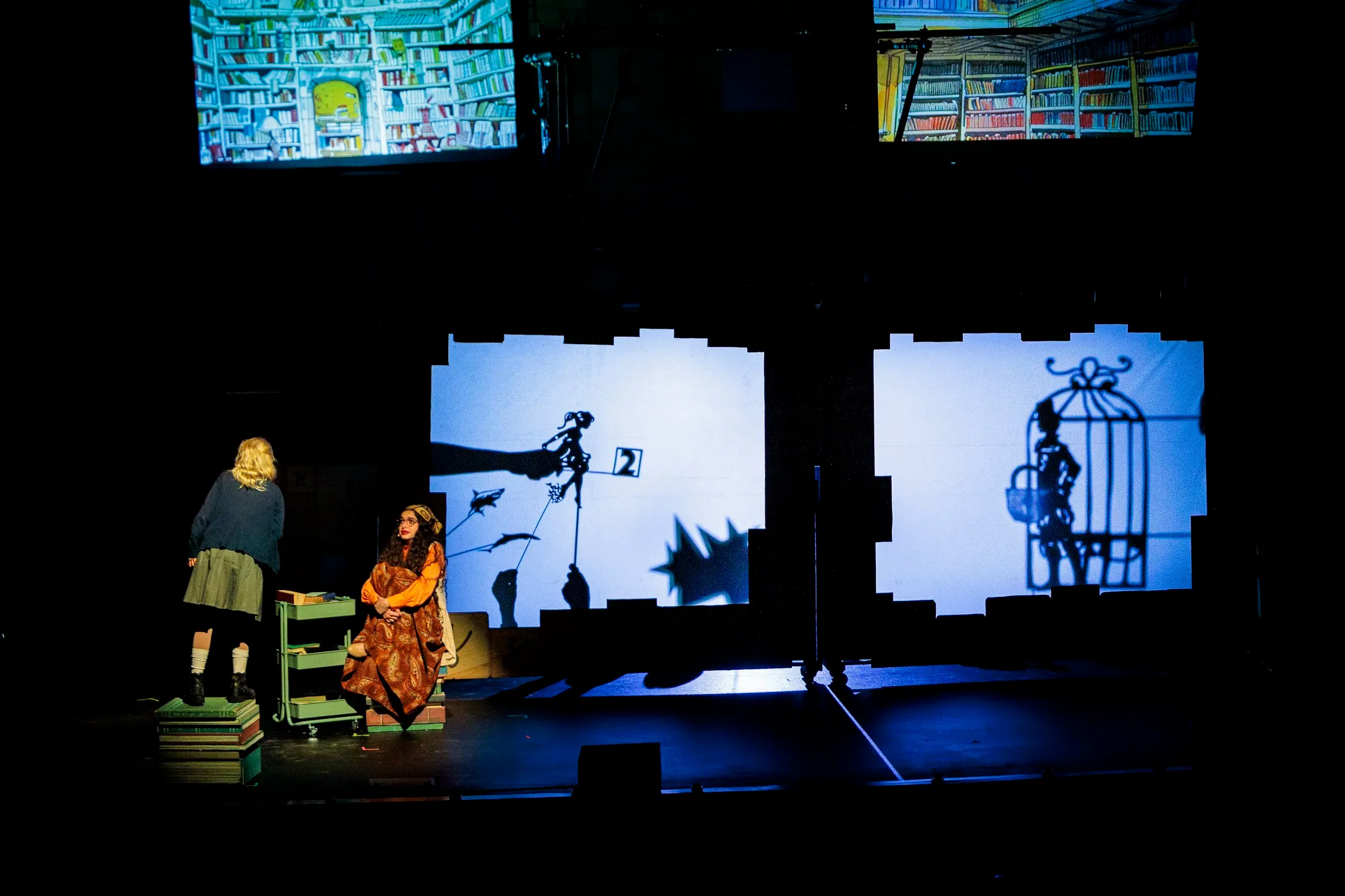 Shadow puppet theater performance with two costumed actors on stage beside illuminated projection screens.