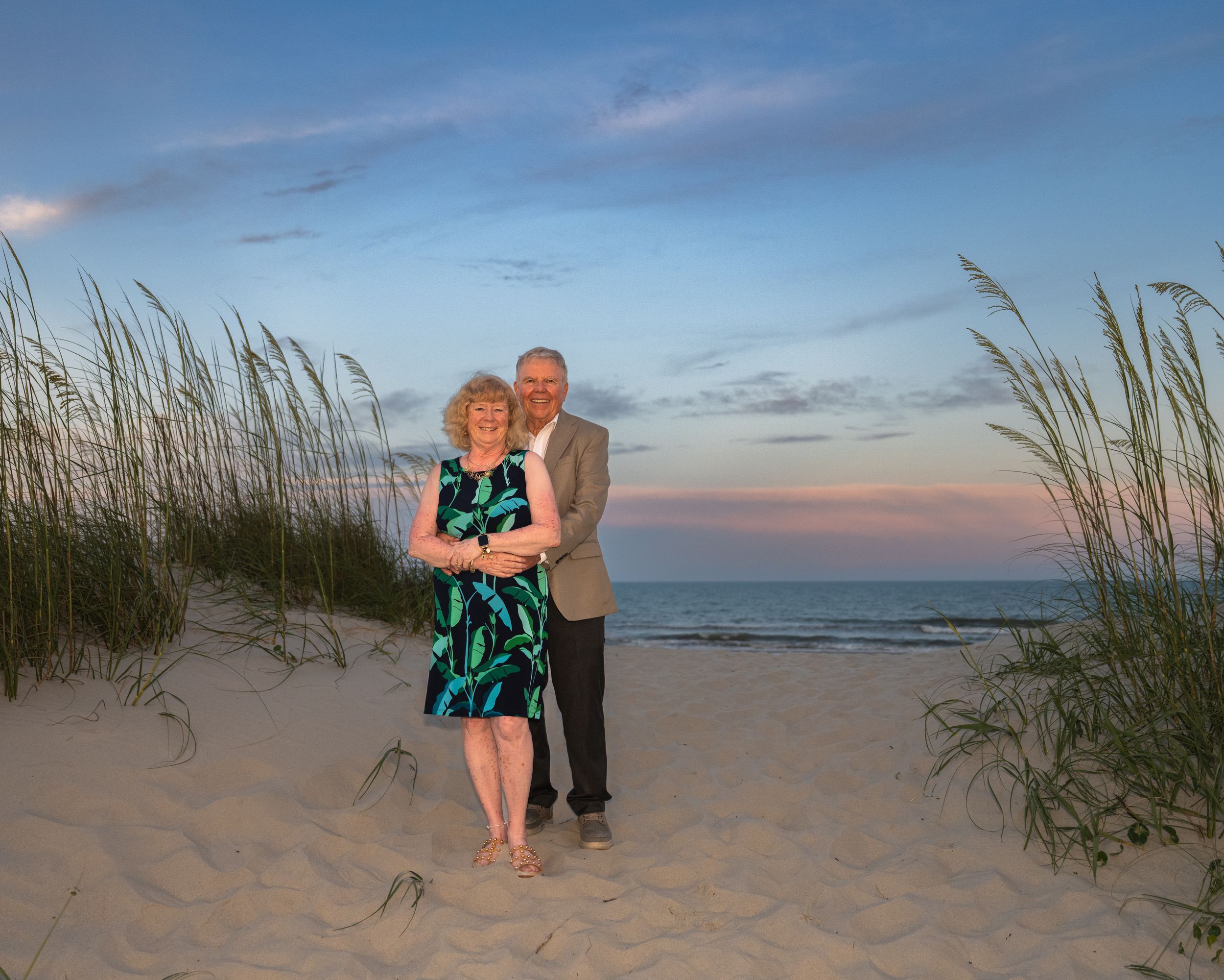 A couple stands together on a sandy beach at sunset with sea oats framing the peaceful coastal scene.