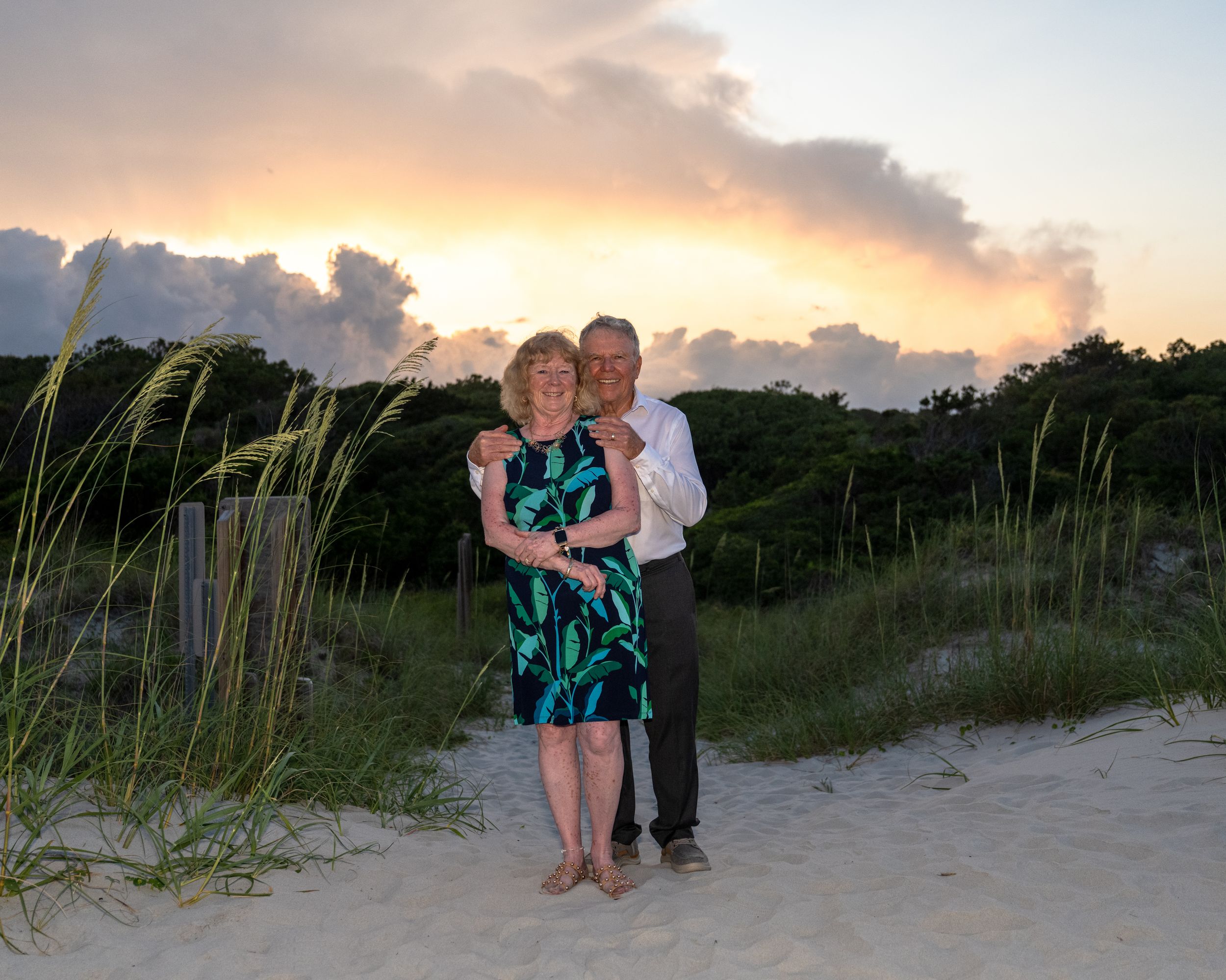 A couple shares a romantic moment on a beach at sunset while standing among sea grass and sand dunes.