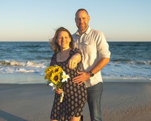 A couple stands together on a sunset beach, with yellow sunflowers in a bouquet and waves rolling in the background.