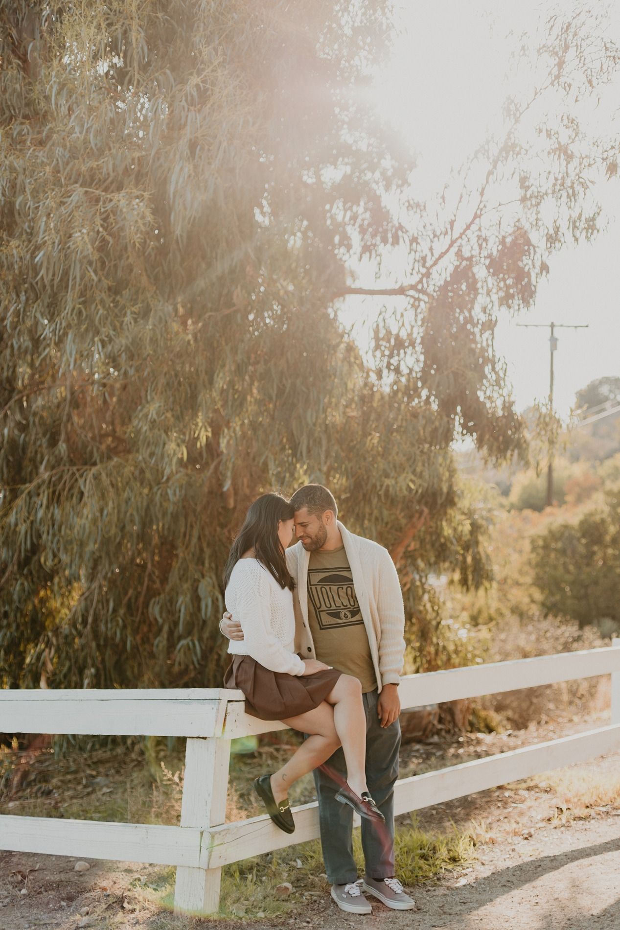 A couple sits together on a white fence along a sunlit path surrounded by willow trees during golden hour at Will Rogers State Park.