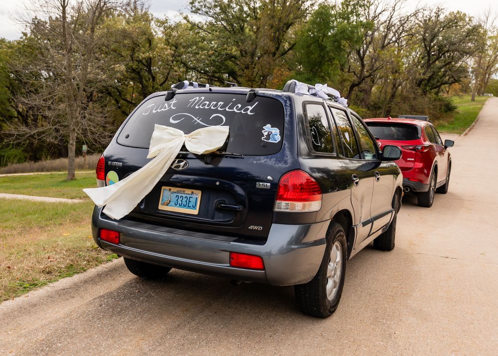 A black SUV decorated with Just Married sign and white bow on bumper parked on a tree-lined road.
