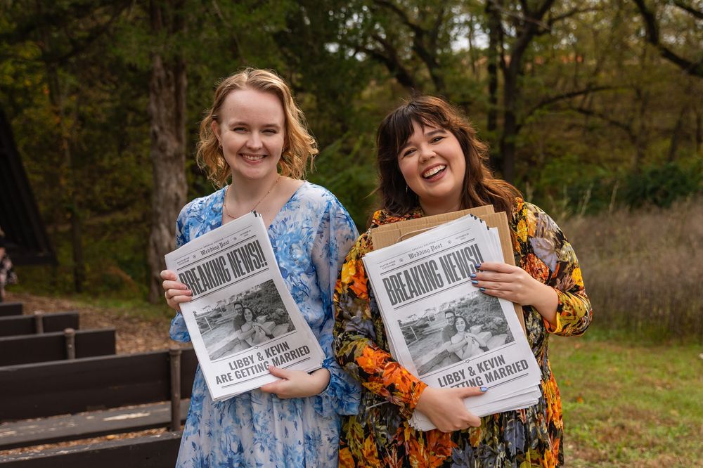 Two women smiling outdoors holding Breaking News newspapers, wearing floral dresses near wooded steps at Platte River State Park